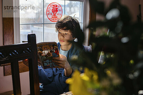 Ein kleiner Junge sitzt am Tisch im Fensterlicht und liest ein Buch