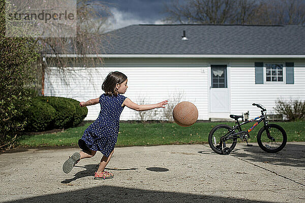 Ein kleines Mädchen jagt in der Einfahrt vor bewölktem Himmel einen Basketball