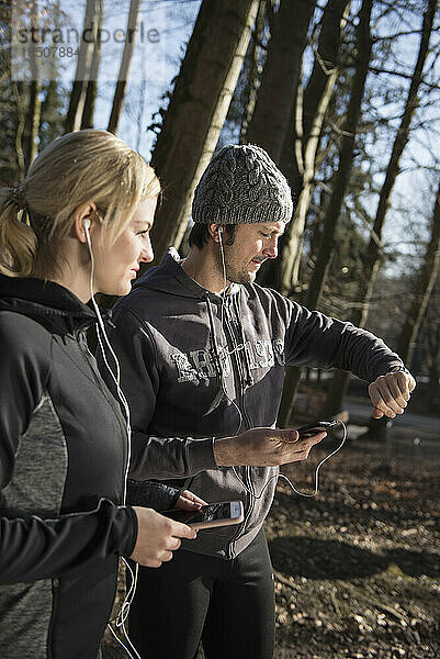 Mann und Frau in Sportkleidung halten Smartphone mit Kopfhörern in der Hand und schauen im Wald auf die Uhrzeit