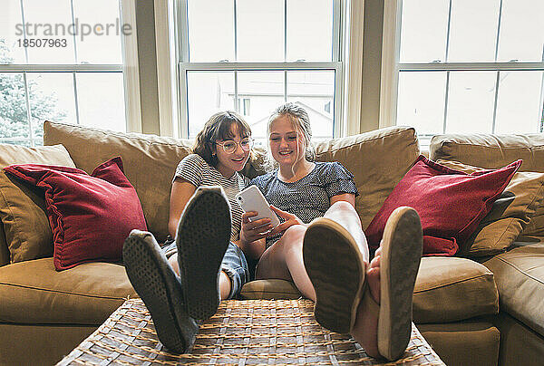 Zwei Mädchen im Teenageralter sitzen mit erhobenen Füßen auf der Couch und schauen auf ihr Handy.