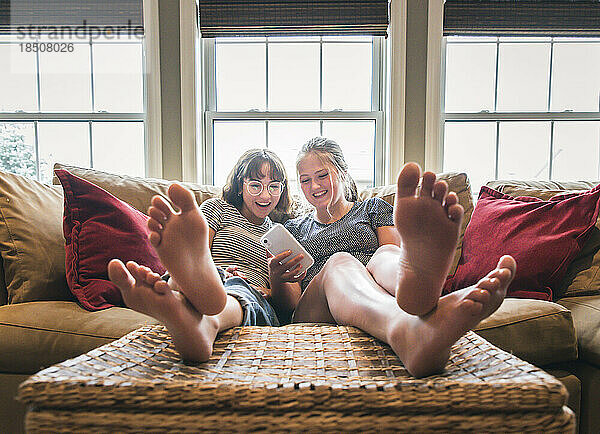 Zwei Mädchen im Teenageralter sitzen mit erhobenen Füßen auf der Couch und schauen auf ihr Handy.