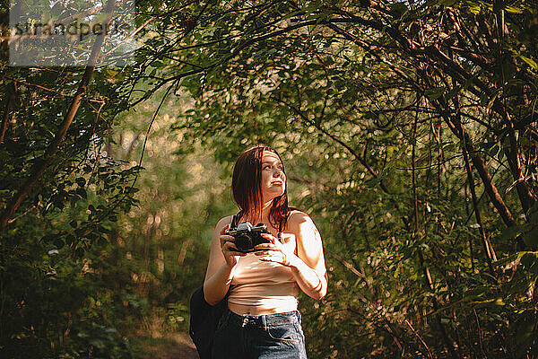 Weibliche Reisende mit Kamera  die wegschaut  während sie im Wald steht