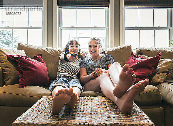 Zwei Mädchen im Teenageralter sitzen mit erhobenen Füßen auf einer Couch und schauen fern.