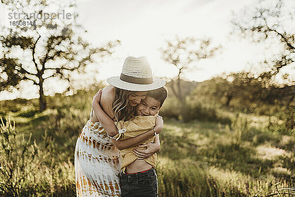 Junge Mutter umarmt Sohn auf sonniger Wiese mit Hut