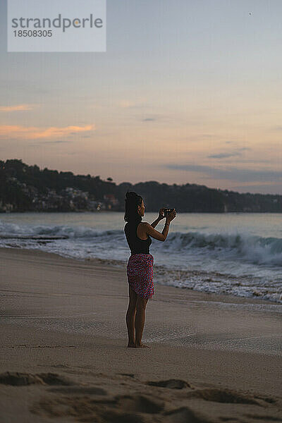 Frau fotografiert mit Kamera einen Sonnenuntergang am Meer.