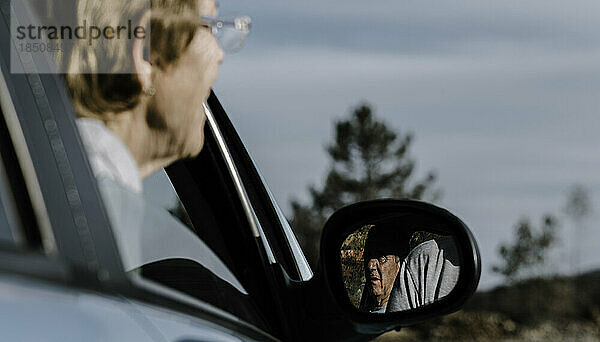 Eine alte Frau ist unscharf  blickt mit dem Kopf aus dem Fenster und konzentriert sich auf den älteren Mann  der sich im Autospiegel spiegelt