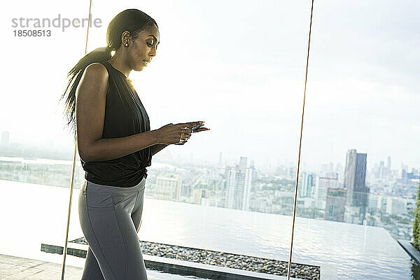 Athletische schwarze Frau benutzt ihr Telefon im Freien mit Panoramablick auf die Stadt.