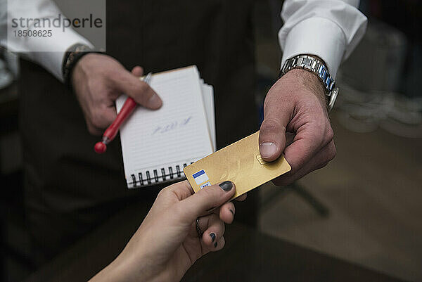 Mann hält Notizblock und Stift in der Hand  während er Kreditkartenzahlung von einer Frau entgegennimmt