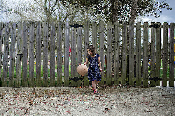 a small girl dribbles a basketball in a driveway in front of fence