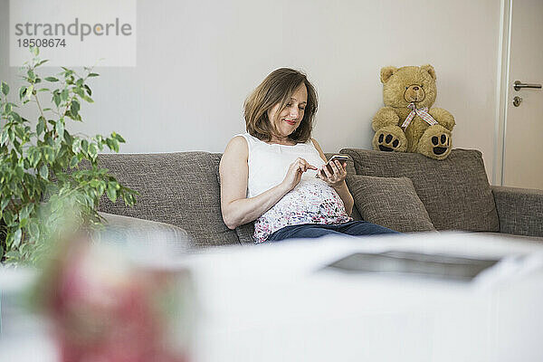 Schwangere Frau sitzt auf dem Sofa und nutzt ihr Mobiltelefon  München  Bayern  Deutschland