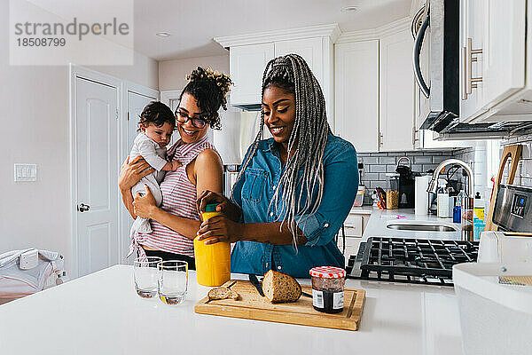 Glückliche Mutter trägt ihren kleinen Jungen  während ihre Freundin in der Küche das Frühstück zubereitet