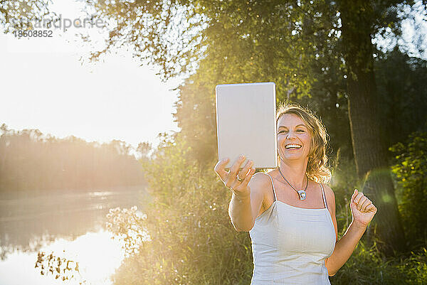 Fröhliche Frau lächelt  während sie ein digitales Tablet am Flussufer benutzt  Bayern  Deutschland