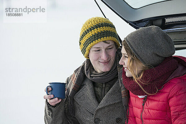 Junges Paar sitzt im Winter mit einer Kaffeetasse auf dem Kofferraum eines Autos  Bayern  Deutschland