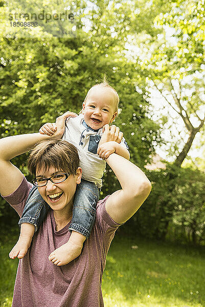Lächelnde Mutter mit ihrem kleinen Sohn auf den Schultern im Garten
