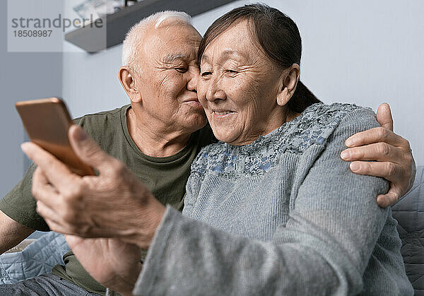 Älteres Paar nutzt Smartphone. Älterer Mann küsst seine Frau