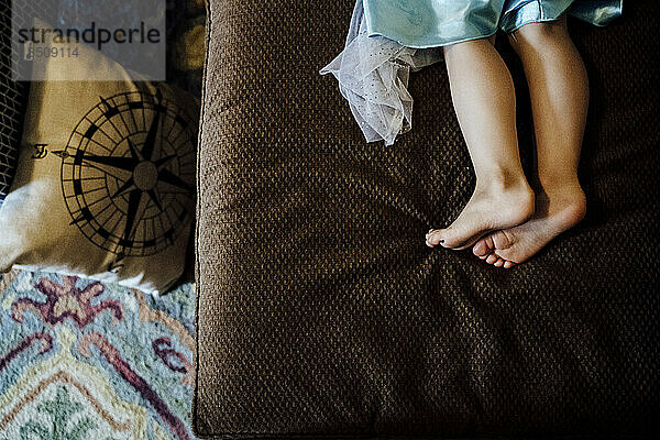 Blick von oben auf die Füße eines jungen Mädchens im Prinzessinnenkleid auf der braunen Couch