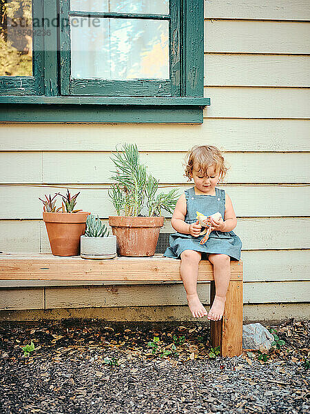Toddler sits on bench with potted plants in farmhouse yard.
