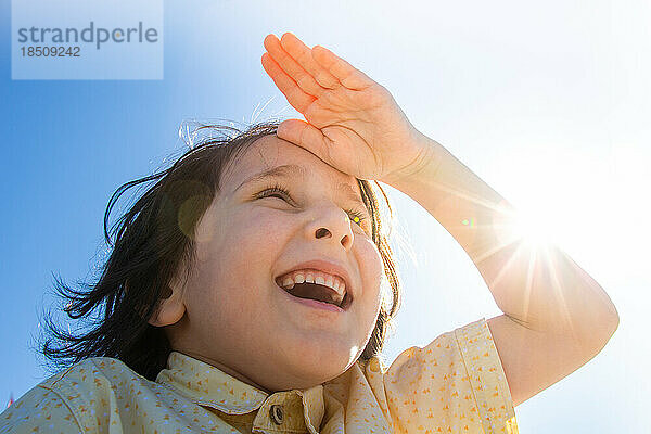 Fröhlicher lächelnder Junge im strahlenden Sonnenschein mit der Hand an der Stirn