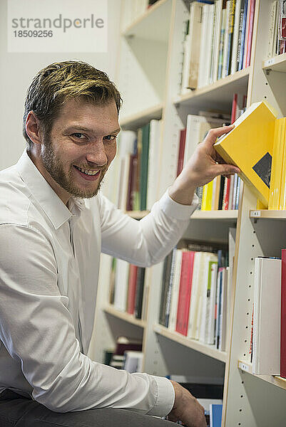 Porträt eines mittleren erwachsenen Geschäftsmannes  der Bücher aus dem Bücherregal in einem Büro in Bayern auswählt