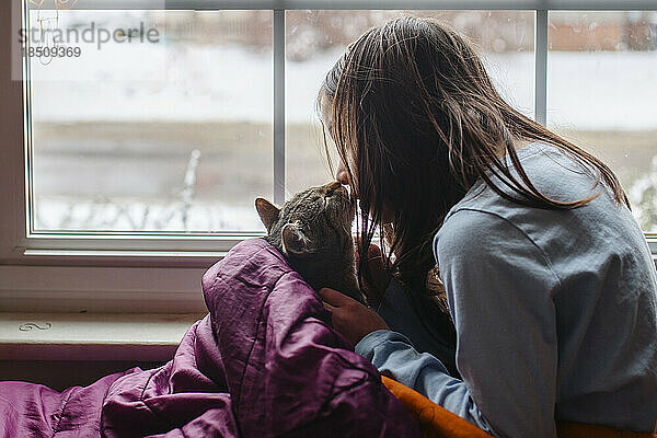 Girl and cat affectionately sit together by window in sleeping bag