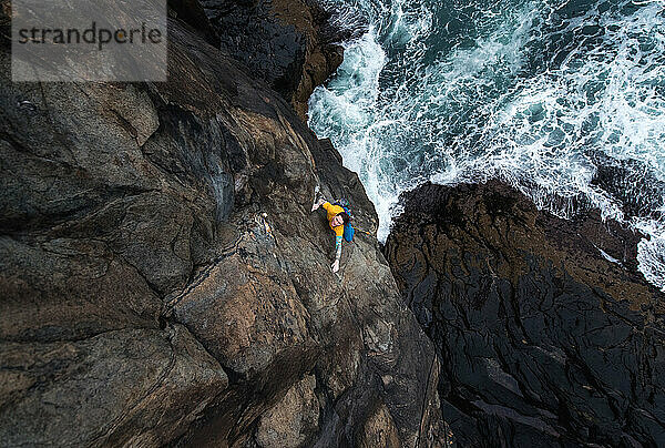 Mann klettert auf einer Klippe am Meer  darunter krachen die Wellen