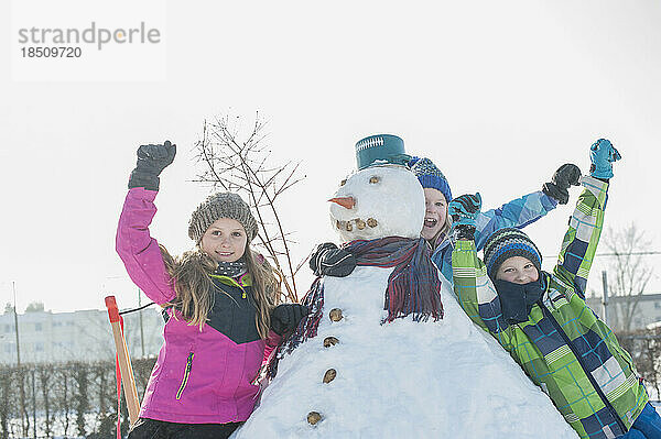 Porträt von Kindern  die sich mit erhobenen Armen auf einen Schneemann stützen