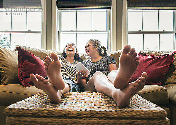 Zwei Mädchen im Teenageralter sitzen mit erhobenen Füßen auf der Couch und lachen.