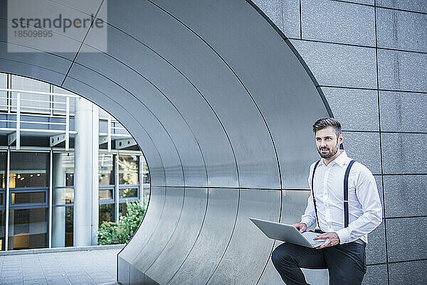 Geschäftsmann lehnt an moderner Architektur und arbeitet am Laptop  München  Bayern  Deutschland