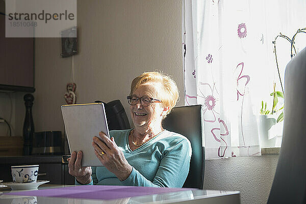Glückliche alte Frau schaut am Esstisch auf ein digitales Tablet