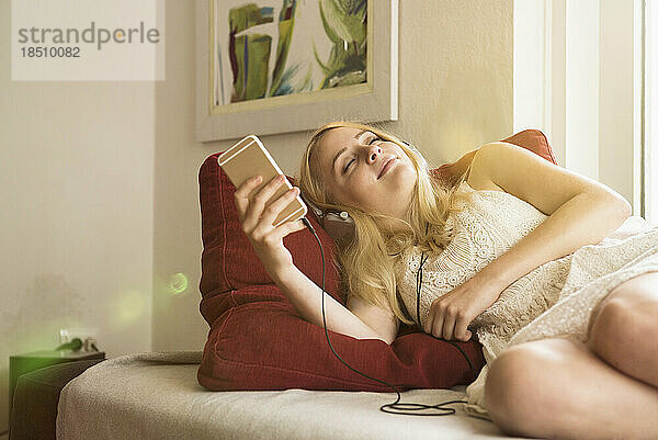 Schöne junge Frau hört Musik und entspannt auf dem Sofa im Wohnzimmer  München  Bayern  Deutschland