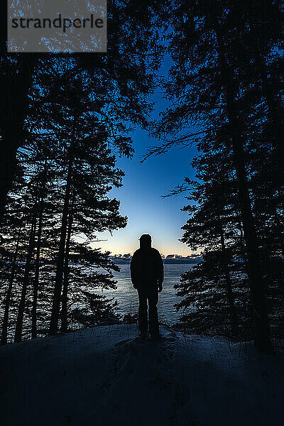 Der Mann steht im Winter im Morgengrauen als Silhouette vor dem Himmel neben dem Meer