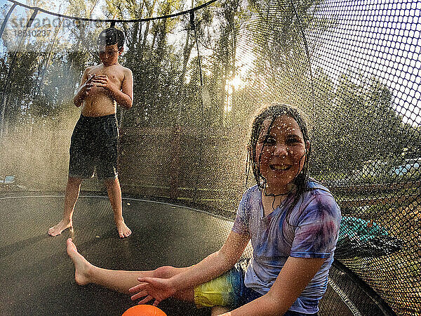 Lächelndes Kind auf Trampolin mit Bruder im Hintergrund und Wasserspray