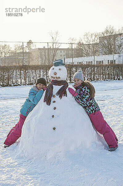 Zwei Mädchen stützen sich auf einen Schneemann auf einem Schneefeld