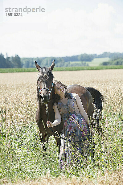 Reife Frau steht mit ihrem braunen Pferd auf einem Weizenfeld und lächelt  Bayern  Deutschland