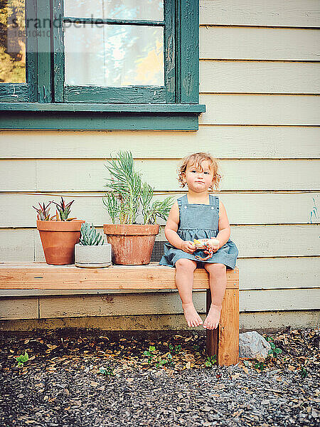Toddler sits on bench with potted plants in farmhouse yard.