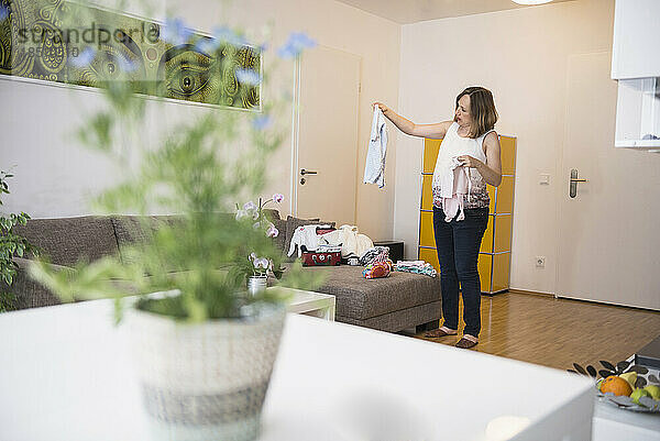 Schwangere Frau steht im Wohnzimmer und schaut sich Babykleidung an  München  Bayern  Deutschland