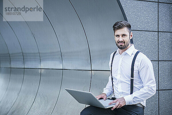 Geschäftsmann lehnt an moderner Architektur und arbeitet am Laptop  München  Bayern  Deutschland