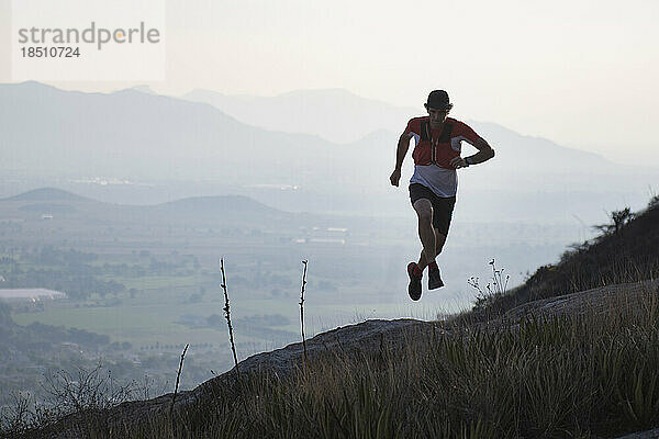 Männlicher Athlet erhebt sich in die Luft  während er in einem Wüstengebiet mit Bergen im Hintergrund in der Nähe von El Arenal  Hidalgo  Mexiko  läuft.