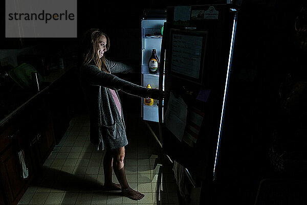 Junge erwachsene Frau schaut in dunkler Küche in offenen Kühlschrank