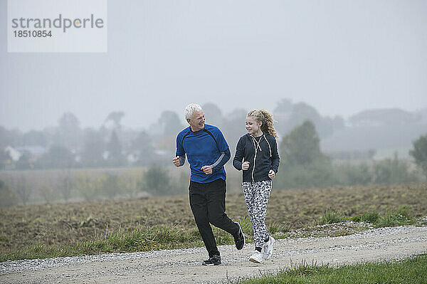 Vater mit seiner Tochter joggt auf Fußweg und lächelt im Morgengrauen  Bayern  Deutschland