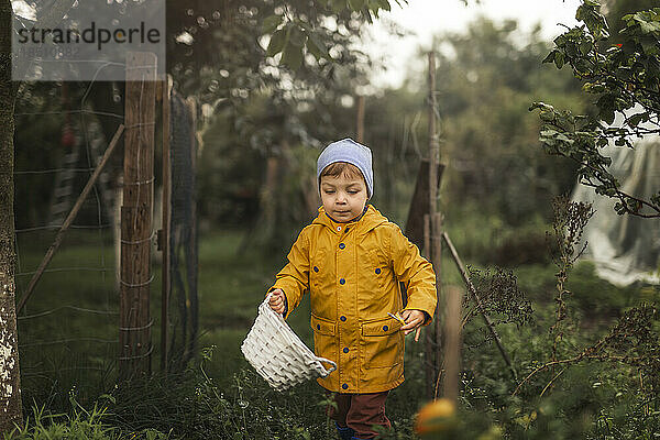 Junge im gelben Regenmantel geht mit weißem Korb und einem Korb durch den Garten