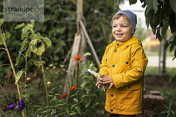 Junge im gelben Regenmantel hält eine Schere im Garten und lächelt