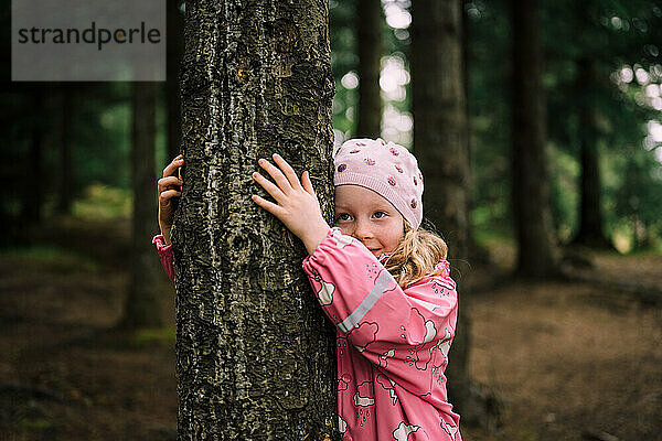 Blondes Kind im Wald lächelt und umarmt Baum