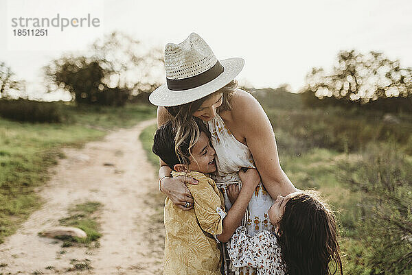 Junge Mutter mit Hut wird von kleinen Kindern auf dem Feld umarmt