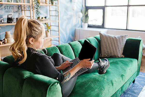 Erwachsene blonde Frau faulenzt auf Samtcouch und hält Laptop in der Hand