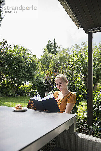 Schöne junge Frau trinkt eine Tasse Tee und liest ein Buch im heimischen Garten  München  Bayern  Deutschland