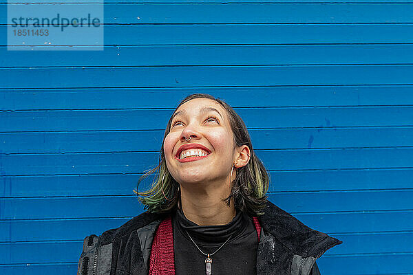 Eine lächelnde Frau blickt auf den Regen  der ihr ins Gesicht fällt  an einer blauen Wand.