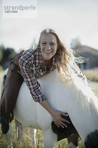 Porträt einer Frau mittleren Alters  die auf dem Rücken eines Pferdes auf einem Bauernhof liegt und lächelt  Bayern  Deutschland