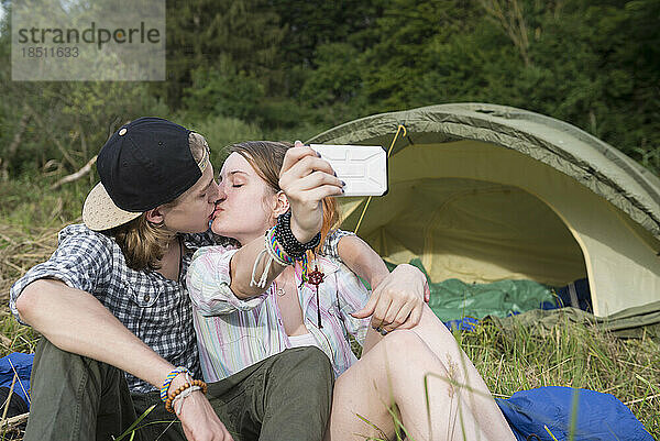 Junges Paar küsst sich und macht ein Selfie mit dem Smartphone vor dem Zeltlager  Bayern  Deutschland