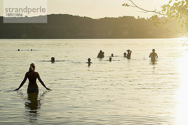 Reife Frau geht bei Sonnenuntergang im See spazieren und ihre Freunde schwimmen  Bayern  Deutschland